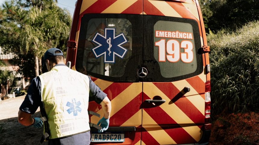 Profissional de emergência ao lado de uma ambulância em uma via urbana brasileira.